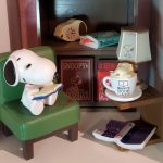 Vintage Peanuts display featuring Snoopy reading a book while seated in a green miniature armchair, set against a wooden bookshelf filled with miniature items like a "Snoopy Book Cafe" mug, desk lamp, and books