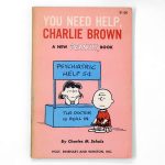 Front cover of the vintage paperback book "You Need Help, Charlie Brown" by Charles M. Schulz with a $1.00 price mark. The cover features a peach background with an illustration of Lucy sitting in her blue psychiatric help booth labeled "Psychiatric Help 5¢ / The Doctor is Real In" while talking to Charlie Brown.