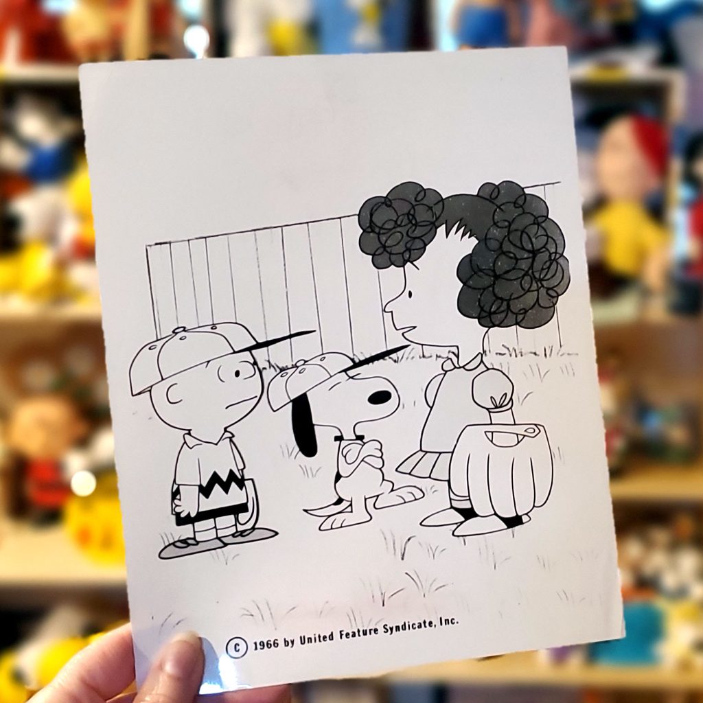 A hand holding a vintage Peanuts comic strip drawing on paper featuring Charlie Brown, Snoopy, and Frieda, with a copyright notice from 1966 by United Feature Syndicate at the bottom.