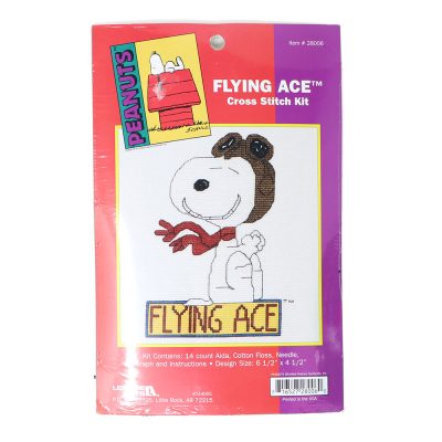 A product package for a Peanuts "Flying Ace" cross-stitch kit by Leisure Arts, featuring an illustration of Snoopy wearing aviator goggles and a red scarf against a purple background, with "FLYING ACE" printed in large letters below.