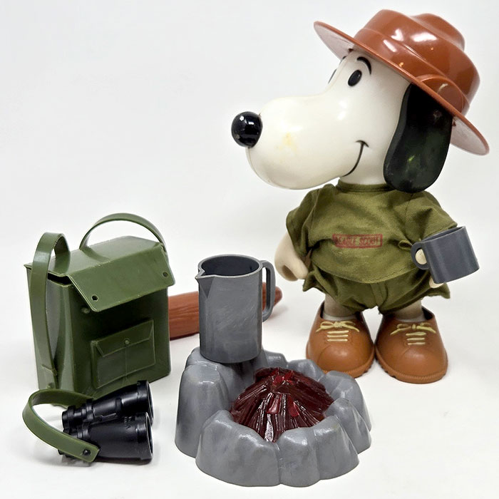 Vintage Snoopy Beagle Scout doll dressed in a brown scout hat, green uniform, and boots, standing next to a matching green backpack, binoculars, and a small campfire set with a coffee pot.