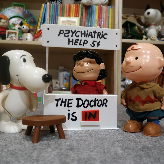 A deluxe Peanuts playset featuring a "Psychiatric Help 5¢" booth with "The Doctor Is In" sign, accompanied by a large Snoopy figure, Lucy sitting behind the counter, and a standing Charlie Brown figure wearing his signature zigzag shirt.