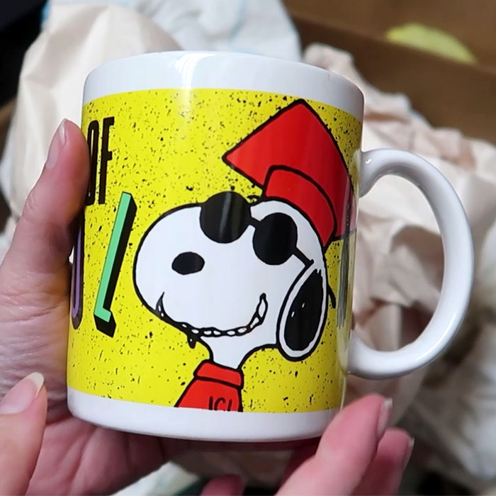 Hand holding a white ceramic Joe Cool Snoopy Mug with a yellow background, featuring a graphic of Snoopy wearing sunglasses, a red cap, and a red shirt with the text "ICU", set against a blurry background of a cardboard box.