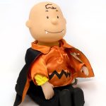 Plush Vampire Charlie Brown doll dressed in his classic yellow-and-black zig-zag shirt with vampire fangs, an orange collar, and a black cape lined with orange, sitting against a white background.