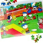 The Baseball Diamond Peanuts Puzzle Build featuring an in-progress jigsaw puzzle depicting a Peanuts baseball game scene with Charlie Brown, Snoopy, and the gang, with a few loose puzzle pieces scattered around the box and main image.