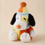 Snoopy Warmies plush stuffed animal sitting on a plain beige background, wearing a bright patchwork clown outfit in orange, yellow, and blue with white paws, an orange nose, a light blue collar, and a yellow and orange party hat topped with a pom-pom.