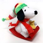 Snoopy wearing a green knit hat and a red scarf while riding on a red sled with Woodstock peeking out from behind him.