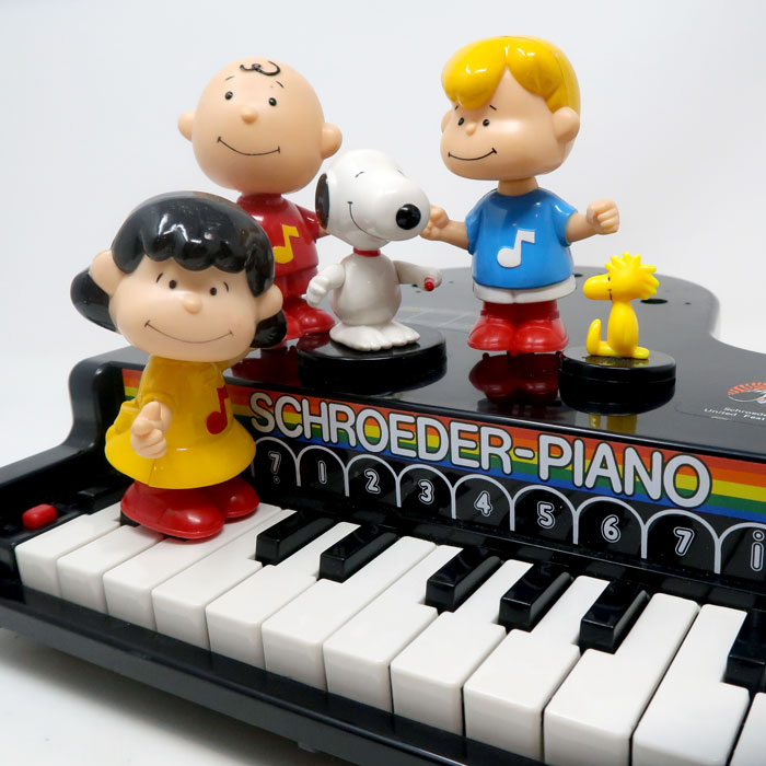 Peanuts toy piano labeled "Schroeder-Piano" with plastic figurines of Charlie Brown, Linus, Lucy playing a mini piano on the keys, Snoopy, Woodstock, and Schroeder arranged on top of it.