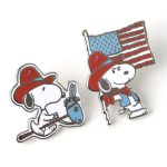 Two enamel pins from Pintrill's Snoopy Yee Haw America collection: one of Snoopy riding a hobby horse wearing a red cowboy hat, and another of Snoopy walking while carrying an American flag and wearing a red hat and vest.
