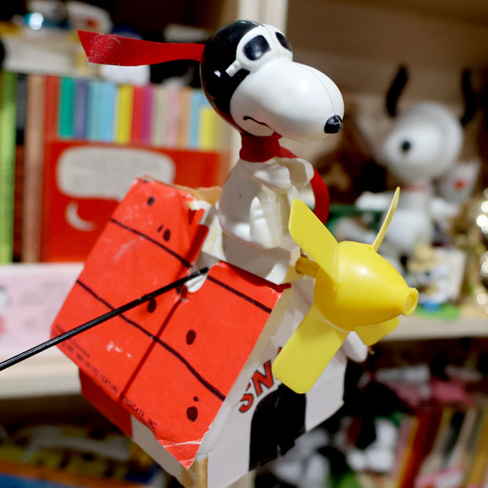 Snoopy wearing an aviator helmet and scarf, sitting atop a toy model of his red flying doghouse with a yellow propeller.