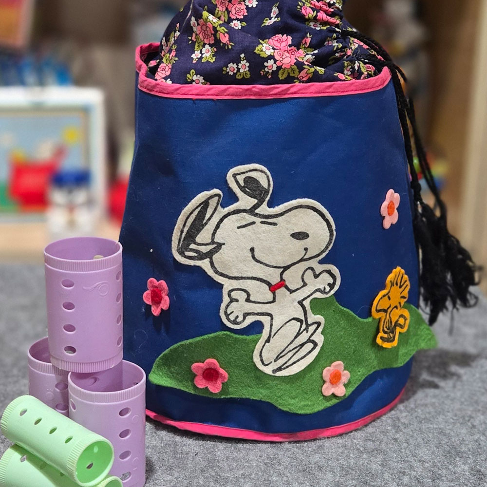 Blue Peanuts drawstring bag featuring a large applique of Snoopy sitting on a green hill with Woodstock, accompanied by pastel purple and green hair rollers.
