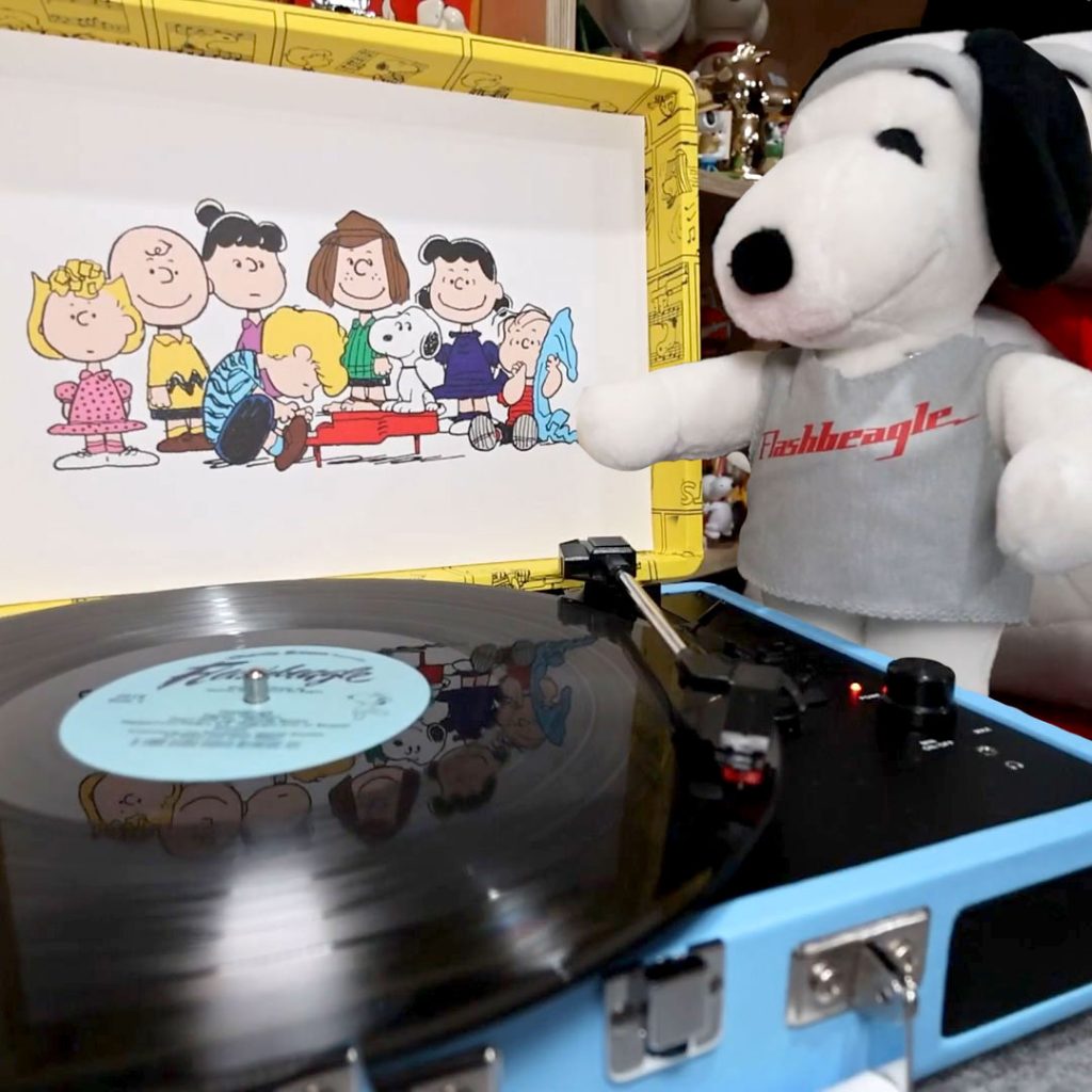 A bright blue Crosley portable briefcase turntable playing a vinyl record, with a large plush Snoopy wearing a gray "Frashbeagle" t-shirt sitting next to it, and the inside lid displaying a graphic of the Peanuts gang.