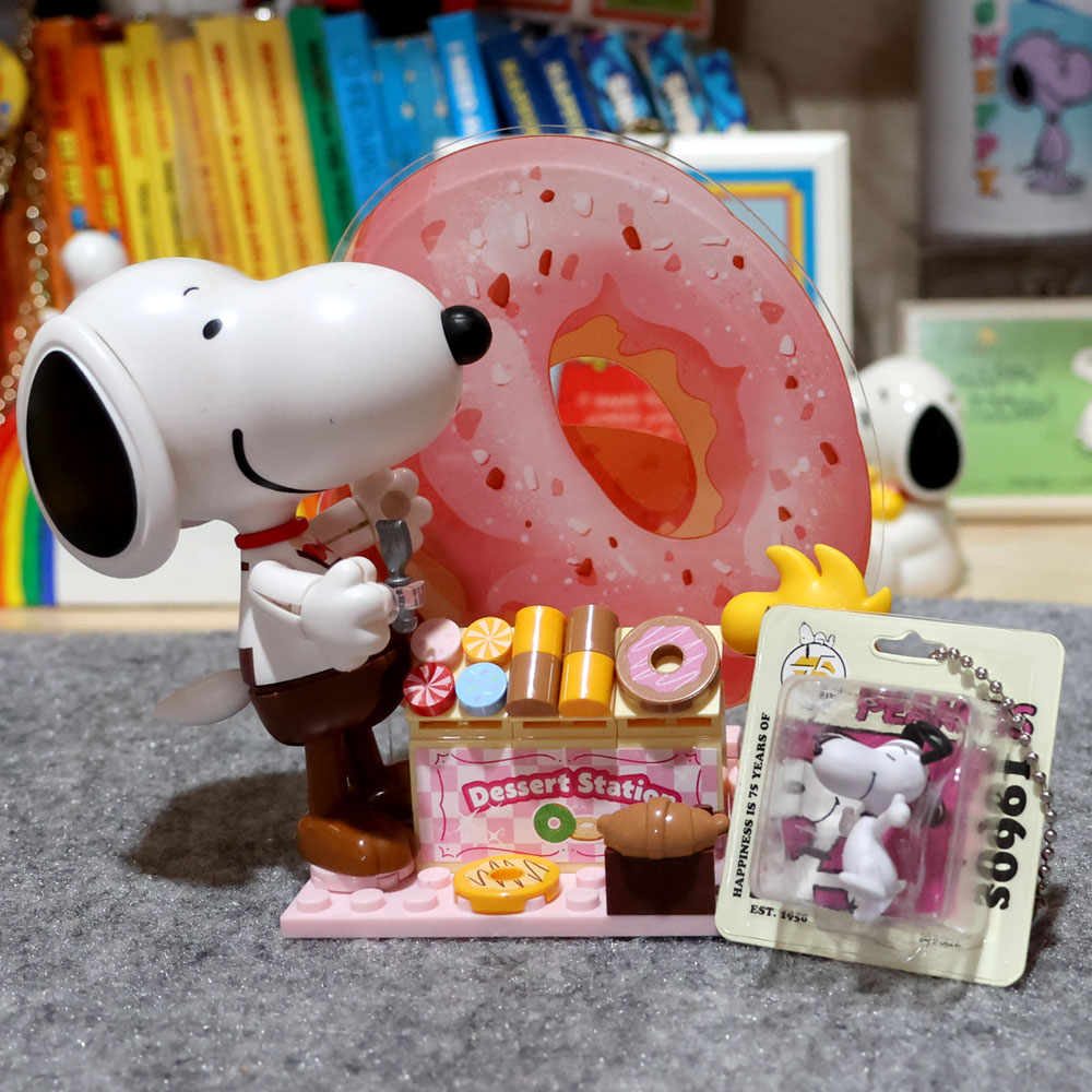 A display of Snoopy Finds at Mitsuwa Marketplace featuring a large translucent pink donut-shaped case, a Snoopy figurine in a chef outfit, a small "Dessert Station" building set, and a packaged 1960s Snoopy charm against a shelf background.