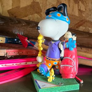 Snoopy dressed in a blue helmet, purple shirt, and orange shorts, holding a yellow object while standing on a green base next to a red bag, with colorful toy skis in the background.