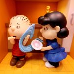DeAgostini Peanuts collectible figurines featuring Linus Van Pelt looking worried while clutching his security blanket and sucking his thumb, standing next to Lucy Van Pelt in her blue dress holding a pink dish, set inside a miniature doghouse room.
