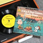 A Charlie Brown Records read-along book titled "It Was A Short Summer, Charlie Brown" and its vinyl record resting on an orange Crosley portable record player, featuring illustrations of Charlie Brown roasting a marshmallow over a campfire and Snoopy holding a toasted marshmallow.