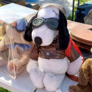Vintage plush Snoopy Flying Ace doll wearing a brown aviator cap, goggles, and a brown leather flight jacket, with another plush toy visible inside a clear display case behind it.