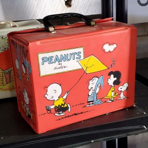 Vintage red Peanuts vinyl lunch box featuring Charlie Brown flying a kite, with Lucy, Linus, Snoopy, and Woodstock on the side graphics and a black plastic handle.