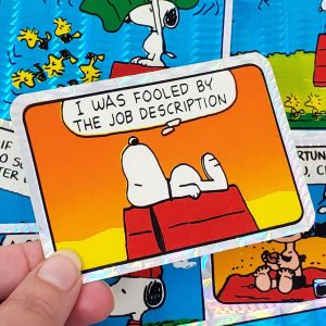 Snoopy prismatic sticker featuring Snoopy lying on his doghouse with a speech bubble that reads "I WAS FOOOLED BY THE JOB DESCRIPTION", held against a background of other comic stickers.