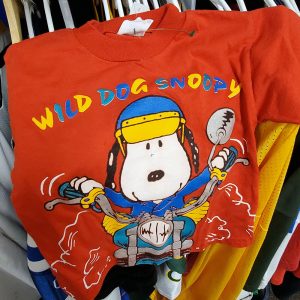 Red "Wild Dog Snoopy" t-shirt featuring Snoopy riding a motorcycle in a yellow helmet and goggles, surrounded by colorful comic graphics and text.