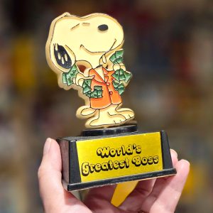 Vintage "World's Greatest Boss" novelty trophy featuring a smiling Snoopy wearing a suit with money sticking out of his pockets, standing atop a black base with a yellow placard.