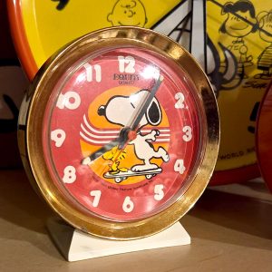Vintage Snoopy skateboarding alarm clock with a round red face, white numerals, and a gold-toned metal frame.