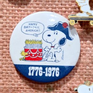 A round vintage Peanuts Bicentennial pinback button featuring Snoopy wearing a tri-fold Revolutionary War hat next to a birthday cake with lighted candles, alongside Woodstock and a speech bubble reading "HAPPY BIRTHDAY, AMERICA!!", with a blue banner at the bottom displaying the dates "1776-1976".