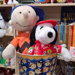 A Charlie Brown plush doll wearing a blue baseball cap and an orange zig-zag shirt, and a Snoopy plush wearing a red baseball uniform, both sitting together inside a decorated Peanuts-themed metal bucket.