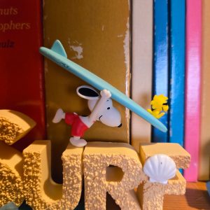 A desk display figurine featuring the word "SURF" in yellow sand-textured 3D block letters with a seashell accent on the letter "F," while Snoopy lies on a blue surfboard attached to the top alongside Woodstock.