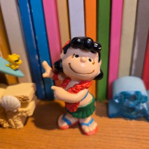 A PVC figurine of Lucy dressed in Hawaiian hula attire, featuring a green grass skirt, red lei, red flower in her hair, and red bikini top, set against a background of colorful books.