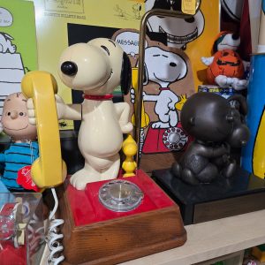 A vintage rotary telephone shaped like Snoopy holding a yellow handset, mounted on a wooden base next to a dark Snoopy statue and surrounded by various Peanuts collectibles.