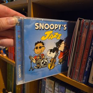 A hand holding up a compact disc jewel case for the album "Snoopy's Jazz", showing illustrations of Schroeder on drums and Snoopy playing double bass wearing sunglasses, set against a background of Peanuts media on a shelf.