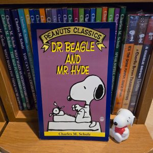 A paperback book titled "Peanuts Classics: Dr. Beagle and Mr. Hyde" by Charles M. Schulz, displaying an illustration of Snoopy typing at his typewriter while perched on top of a giant Snoopy head, set against a bookshelf filled with Peanuts books.