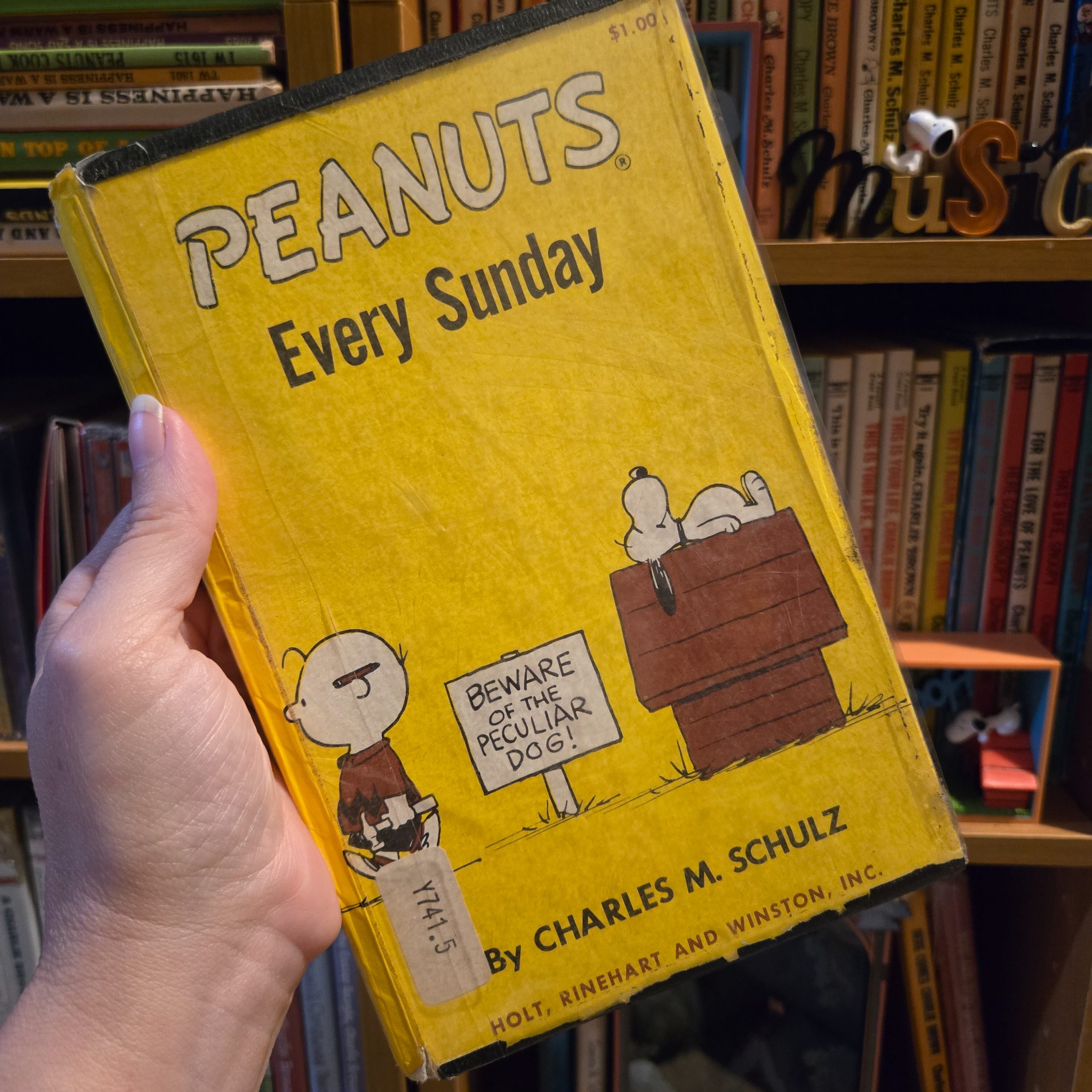 A hand holding a vintage yellow library-bound edition of the Peanuts book "Peanuts Every Sunday" by Charles M. Schulz, published by Holt, Rinehart and Winston, set against a background of bookshelves filled with Peanuts collectibles.
