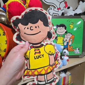 A hand holding a handmade fabric Lucy Van Pelt Stuff N' Lace doll stitched along the edges with yarn, wearing a yellow top with "LUCY" printed in blue text alongside red accents and a pleated skirt, set against a background of Peanuts collectibles.
