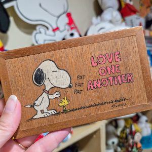 A rectangular wooden Peanuts plaque held in a hand, featuring Snoopy gently patting Woodstock on the head with sound effects "PAT PAT PAT", alongside the text "LOVE ONE ANOTHER" in textured red and pink block letters.