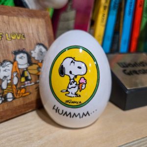 A decorative white ceramic egg featuring an illustration of Snoopy holding a pink ice cream cone against a yellow circular background bordered in green, with Schulz's signature below and the text "HUMMM..." printed near the bottom.