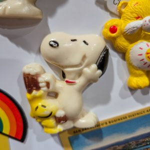 A vintage Peanuts magnet featuring Snoopy holding a glass mug of root beer with Woodstock perched happily in front of him, shown among other collectible items.