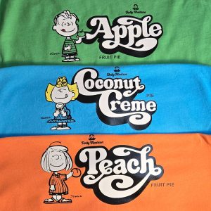 Three vintage Dolly Madison Fruit Pie T-shirts stacked on top of each other in green, blue, and orange, featuring Linus next to "Apple," Sally next to "Coconut Creme," and Peppermint Patty next to "Peach."
