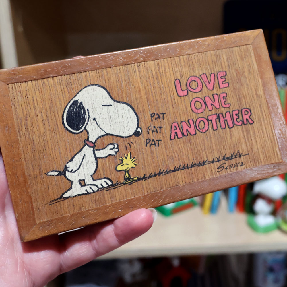 A hand holding a wooden Hallmark miniature gallery plaque featuring Snoopy gently patting Woodstock on the head with "PAT PAT PAT" sound effects, next to the inscription "LOVE ONE ANOTHER" in pink block lettering.