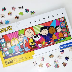 A Peanuts 1000-piece panorama jigsaw puzzle by Clementoni partially assembled on a white surface, surrounded by scattered puzzle pieces and showing the completed box art featuring the Peanuts gang.