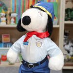Plush Snoopy doll dressed in a vintage police officer uniform. He wears a light blue button-up shirt featuring a "Snoopy" name tag and a "Police Officer" badge patch, a red ribbon neckerchief, navy blue trousers with a black buckle belt, and a matching navy police hat with yellow trim.