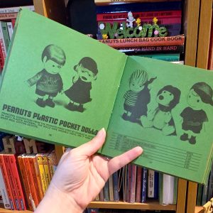 Open green book spread of Peanuts Plastic Pocket Dolls catalog, showing illustrations of Peanuts characters including Schroeder and Lucy on the left page, and Linus, Snoopy, and Charlie Brown on the right page, with text that reads: "Peanuts Plastic Pocket Dolls... principal characters from the peanuts comic strip by Charles M. Schulz in soft plastic material."
