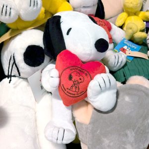 Snoopy plush toy resting among a pile of other plush items and toys, featuring Snoopy's classic black and white design with floppy black ears, embroidered facial features, whiskers, and holding a small red heart.
