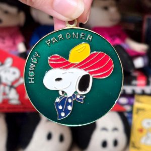 Round green enamel pendant featuring Snoopy wearing a red cowboy hat with a yellow band and a blue patterned bandana, with gold lettering along the top and left side that reads "HOWDY PARDNER", held up against a blurred background with plush toys.