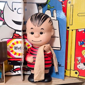 Linus van Pelt plastic Hungerford doll holding his signature security blanket, positioned in front of a Charlie Brown display and a colorful blue-and-yellow background.