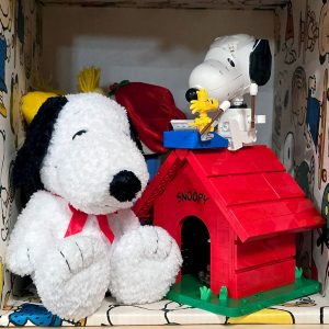 A fluffy white Snoopy plush toy sitting next to a red LEGO doghouse, on top of which a LEGO Snoopy figure and a yellow Woodstock figure are seated, all positioned inside a display shelf lined with Peanuts comic strip wallpaper.