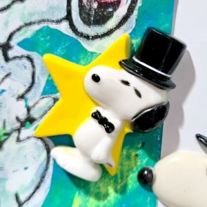 A vintage Snoopy magnet wearing a black top hat and bowtie, posed in front of a bright yellow star against a colorful painted background with a partial view of another Snoopy figure.