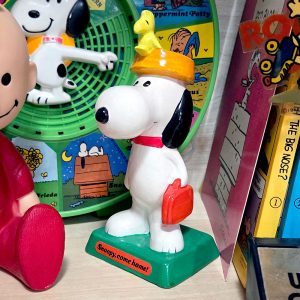 A vintage figurine of Snoopy wearing a yellow dogdish with Woodstock perched on top, holding a red suitcase and standing on a green base that reads "Snoopy, come home!", set among a collection of other Peanuts merchandise.