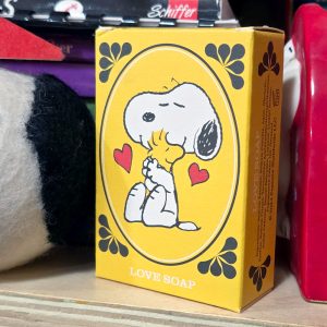 A yellow box of Peanuts "Love Soap" featuring an illustration of Snoopy hugging Woodstock against a yellow oval with red hearts, set among other Peanuts merchandise.